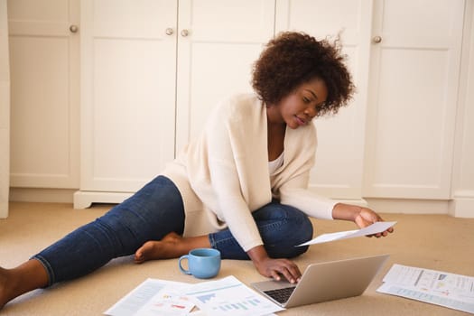 Happy african american woman working in bedroom, sitting on floor using laptop and holding paperwork. flexible working from home.