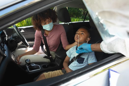 Medical worker wearing ppe suit taking swab tests of african american mother with son sitting in car. medical precautions, lifestyle during covid 19 pandemic.