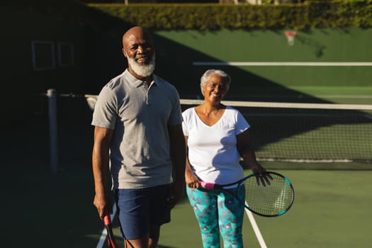 Portrait of smiling senior african american couple with tennis rackets on tennis court. retirement and active senior lifestyle concept.