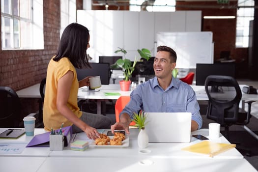 Happy caucasian male and female colleague eating pastries and talking sitting in office. working in business at a modern office.