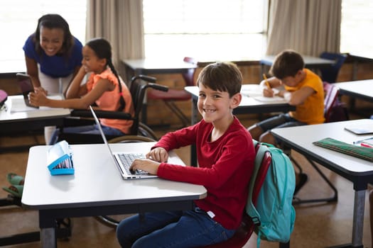 Portrait of caucasian boy with laptop smiling while sitting on his desk at elementary school. school and education concept