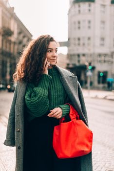 A woman on the street uses a mobile phone. online shopping. use of mobile applications. beautiful young woman with long curly dark hair in a casual coat, trendy green sweater and red handbag