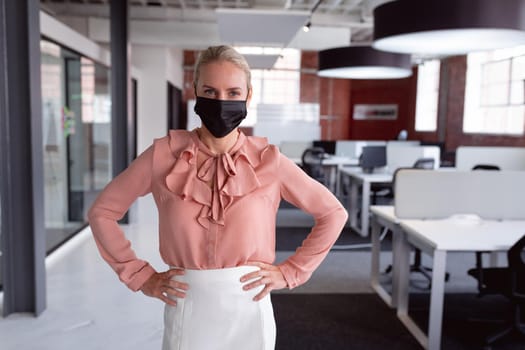 Portrait of caucasian businesswoman in face mask standing in office with hands on hips. working in business at a modern office during coronavirus covid 19 pandemic.