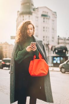 A woman on the street uses a mobile phone. online shopping. use of mobile applications. beautiful young woman with long curly dark hair in a casual coat, trendy green sweater and red handbag