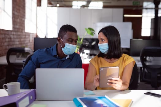 Diverse male and female colleague in face masks sitting at desk discussing, using laptop and tablet. working in business at a modern office during coronavirus covid 19 pandemic.