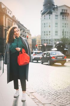 A woman on the street uses a mobile phone. online shopping. use of mobile applications. beautiful young woman with long curly dark hair in a casual coat, trendy green sweater and red handbag