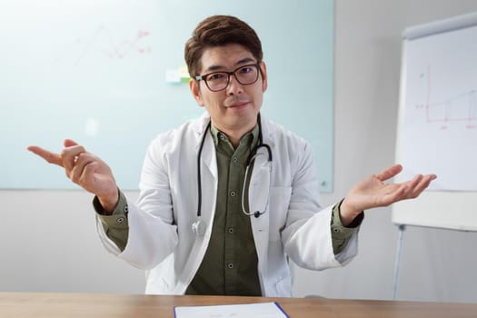 Asian male doctor sitting at desk in office gesturing during video call. telemedicine, online medical and healthcare services during coronavirus covid 19 pandemic.