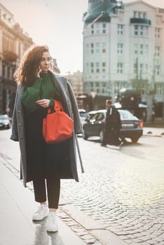 A woman on the street uses a mobile phone. online shopping. use of mobile applications. beautiful young woman with long curly dark hair in a casual coat, trendy green sweater and red handbag