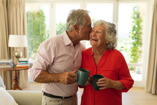 Senior caucasian couple kissing on cheek smiling and holding mugs in living room. retreat, retirement and happy senior lifestyle concept.