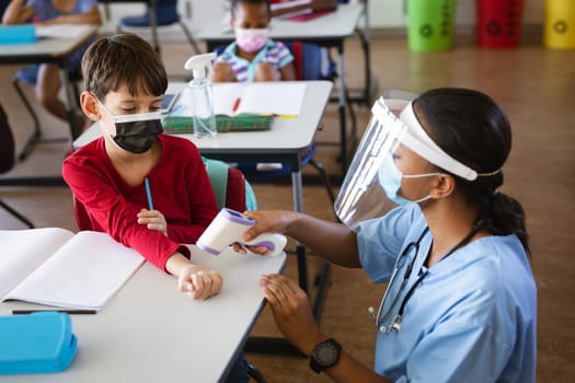 Female health worker wearing face shield measuring temperature of a boy at elementary school. education back to school health safety during covid19 coronavirus pandemic.