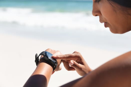 Mixed race woman exercising on beach checking smartwatch. healthy outdoor leisure time by the sea.
