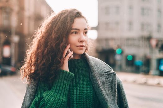 A woman on the street uses a mobile phone. online shopping. use of mobile applications. beautiful young woman with long curly dark hair in a casual coat, trendy green sweater and red handbag