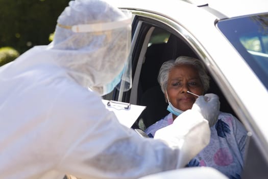 Medical worker wearing ppe suit taking swab test of senior african american woman sitting in car. retirement and senior lifestyle during covid 19 concept.