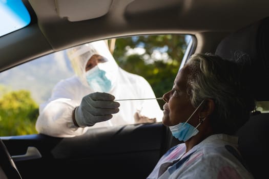 Medical worker wearing ppe suit taking swab test of senior african american woman sitting in car. retirement and senior lifestyle during covid 19 concept.