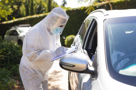 Medical worker wearing ppe suit taking notes talking to senior african american woman sitting in car. retirement and senior lifestyle during covid 19 concept.
