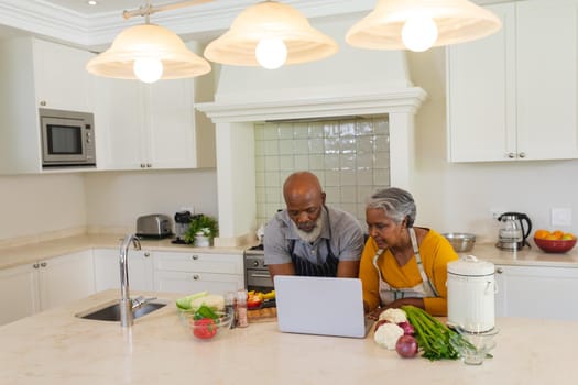 Senior african american couple cooking together in kitchen using laptop. retreat, retirement and happy senior lifestyle concept.