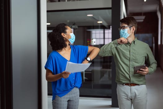 Diverse male and female colleague in face masks greeting by bumping elbows in office corridor. working in business at a modern office during coronavirus covid 19 pandemic.