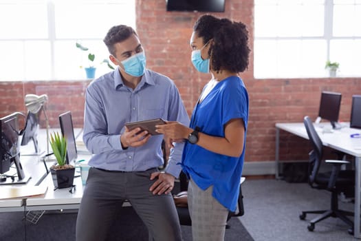 Diverse male and female colleague in face masks looking at tablet and discussing standing in office. working in business at a modern office during coronavirus covid 19 pandemic. .