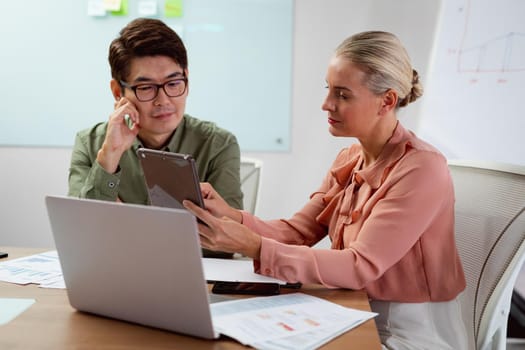 Diverse male and female colleague sitting at table with tablet, laptop and paperwork discussing. working in business at a modern office.