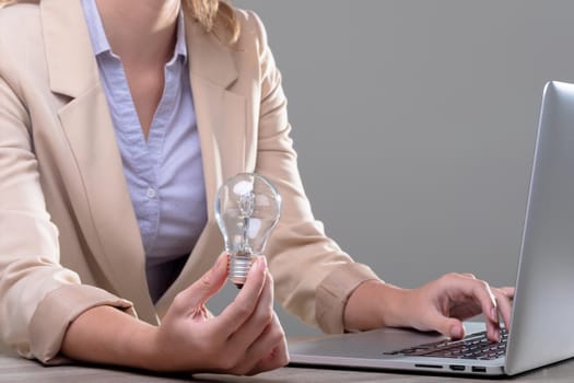 Midsection of caucasian businesswoman holding light bulb using laptop, isolated on grey background. business, technology, communication and growth concept.