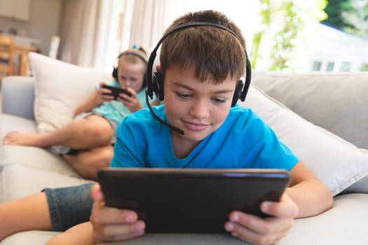 Caucasian brother and sister wearing headphones and using tablets at home. childhood with technology, spending free time at home.