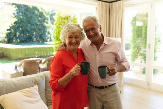 Portrait of senior caucasian couple looking at camera, smiling and holding mugs in living room. retreat, retirement and happy senior lifestyle concept.