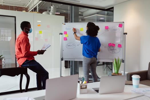 Diverse male and female colleague in face masks brainstorming at whiteboard in office. working in business at a modern office during coronavirus covid 19 pandemic.