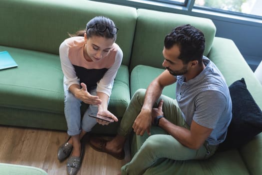 Two diverse creative colleagues sitting on sofa using smartphone. modern office of a creative design business.