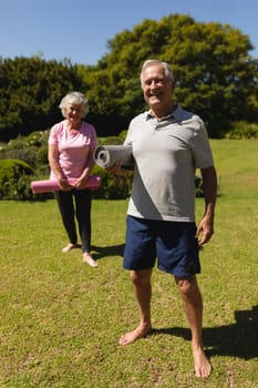 Portrait of senior caucasian couple holding yoga mats, looking at camera and smiling in sunny garden. retirement retreat and active senior lifestyle concept.