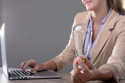 Smiling caucasian businesswoman holding light bulb using laptop, isolated on grey background. business, technology, communication and growth concept.