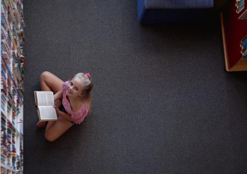 Overhead view of smiling caucasian schoolgirl sitting on floor reading book in school library. childhood and education at elementary school.