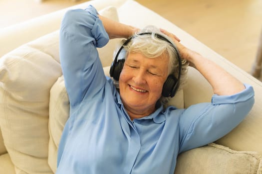 Senior caucasian woman sitting on sofa wearing headphones with eyes closed. retreat, retirement and happy senior lifestyle concept.