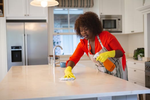 Happy african american woman in kitchen wearing rubber gloves, cleaning kitchen worktop. spending free time at home.