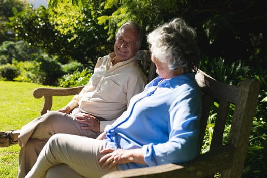 Senior caucasian couple sitting on bench together in sunny garden. retreat, retirement and happy senior lifestyle concept.