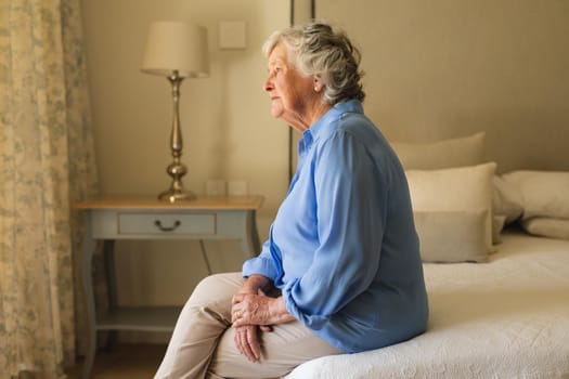 Senior caucasian woman sitting on bed and thinking in bedroom. retreat, retirement and senior lifestyle concept.