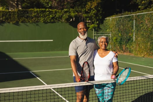 Portrait of smiling senior african american couple with tennis rackets on tennis court. retirement and active senior lifestyle concept.