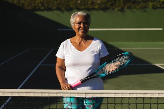 Portrait of smiling senior african american woman holding tennis racket on tennis court. retirement and active senior lifestyle concept.