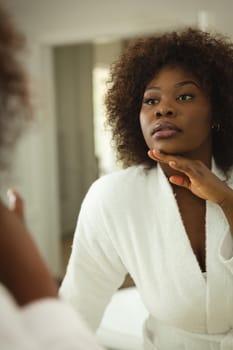 African american woman in bathroom wearing bathrobe, looking in mirror and moisturising face. health, beauty and wellbeing, spending quality time at home.