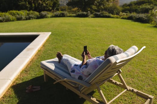 Senior african american woman using smartphone in deckchair by swimming pool in sunny garden. retreat, retirement and happy senior lifestyle concept.