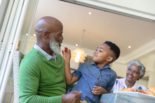 Happy african american grandson and grandparents sitting at table talking during breakfast. family enjoying quality free time together.