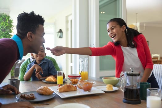 Happy african american son and father sitting at table during breakfast, daughter feeding him. family enjoying quality free time together.