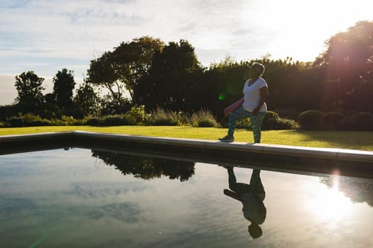 Senior african american woman standing with yoga mat by outdoor pool in countryside. retirement and active senior lifestyle concept.