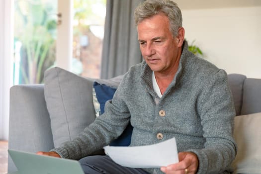 Focused caucasian senior man sitting on sofa, doing paperwork, using laptop. active retirement lifestyle at home.