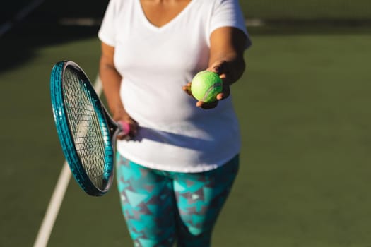 Midsection of senior african american woman holding tennis racket and ball on tennis court. retirement and active senior lifestyle concept.