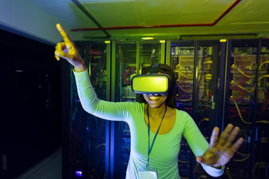 Happy african american female computer technician wearing vr headset and working in server room. digital information storage and communication network technology.