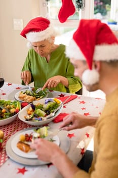 Mother in law and son in law wearing santa hats sitting at christmas table and eating. family christmas time and festivity together at home.