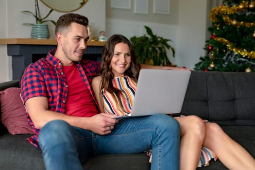 Happy caucasian couple sitting on sofa and having video call on laptop at christmas time. christmas, festivity and communication technology.