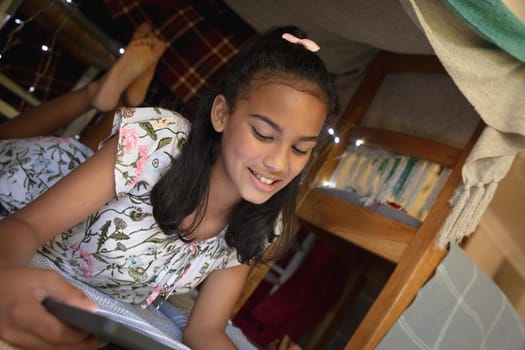 Happy african american girl at home, lying in blanket fort and using tablet. childhood with technology, spending free time at home.