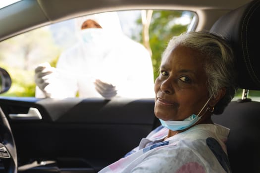 Medical worker wearing ppe suit taking swab test of senior african american woman sitting in car. retirement and senior lifestyle during covid 19 concept.