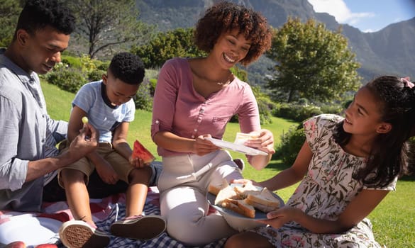 Happy african american couple with son and daughter outdoors, having picnic in sunny garden. family enjoying quality free time together.